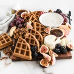 fall brunch board with waffles, yogurt, fruit and cheese on a white marble background.