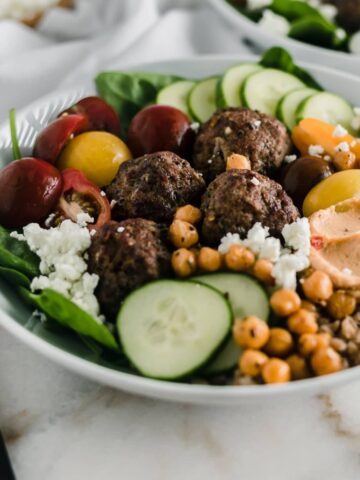 meatballs, cucumbers, hummus, feta and vegetables in a white bowl.