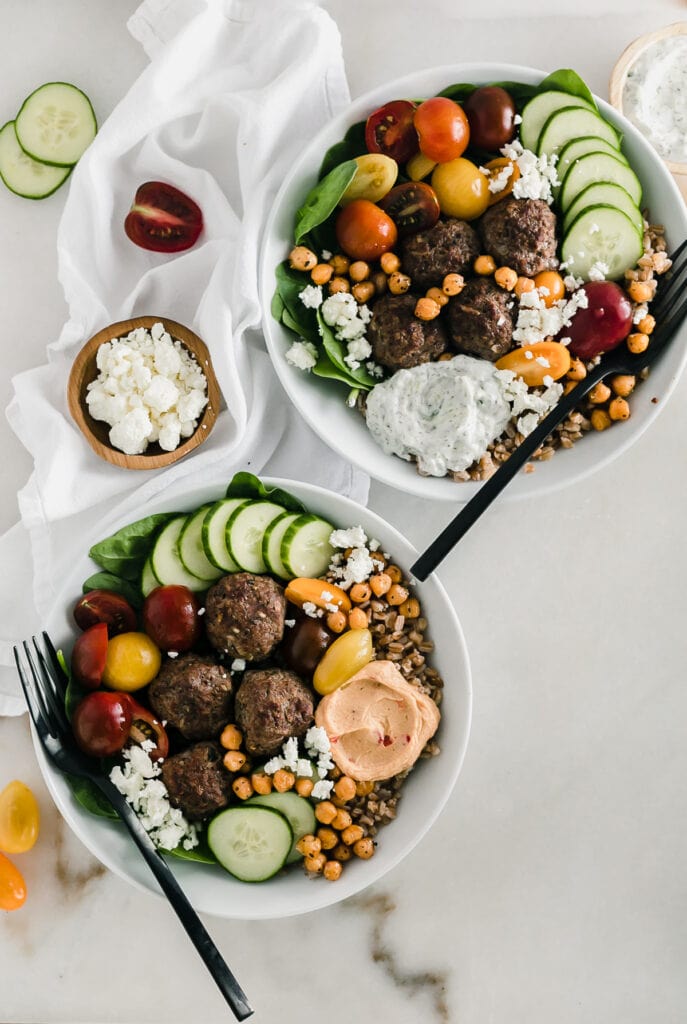 Overhead view of two mediterranean meatball bowls with black forks on a white background.