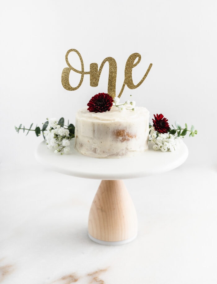 white smash cake on a white stand, topped with flowers and a one cake topper.