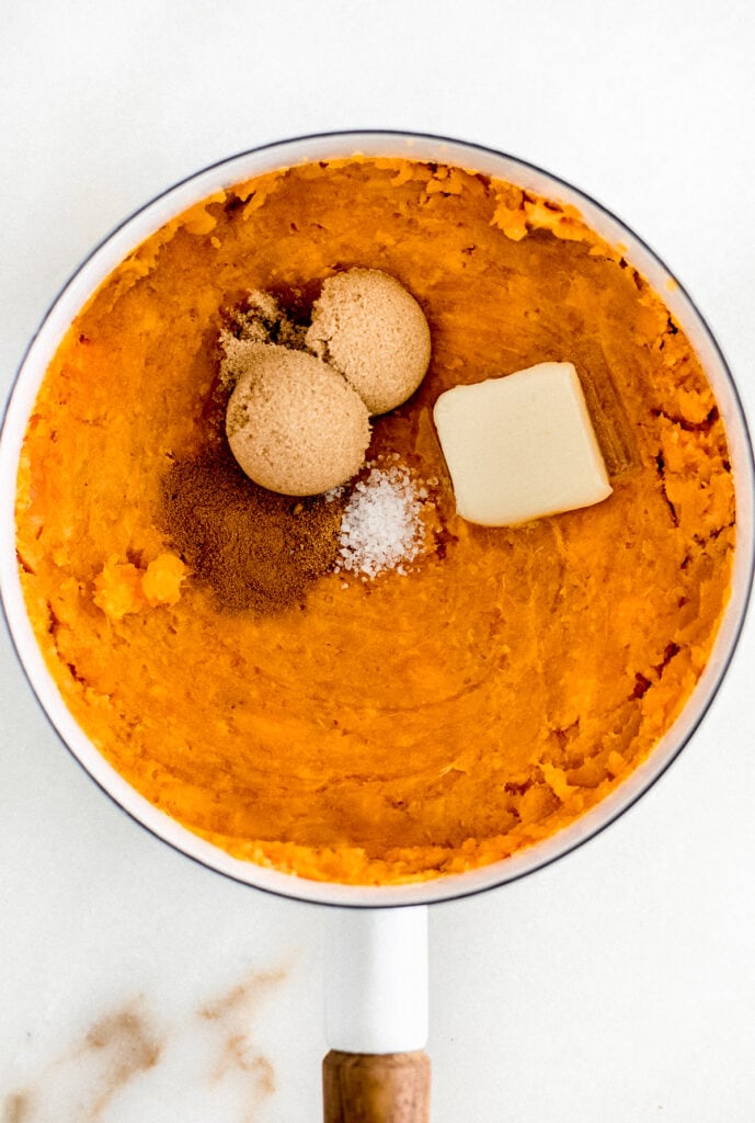 mashed sweet potatoes in a white saucepan with brown sugar, spices and butter on top.