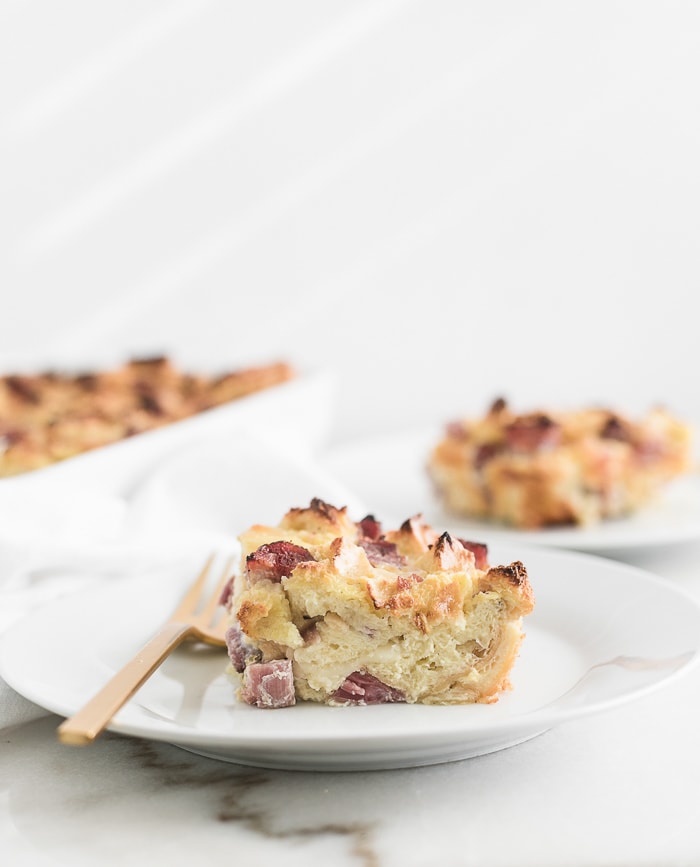 slice of ham and swiss strata on a white plate with a gold fork next to it.