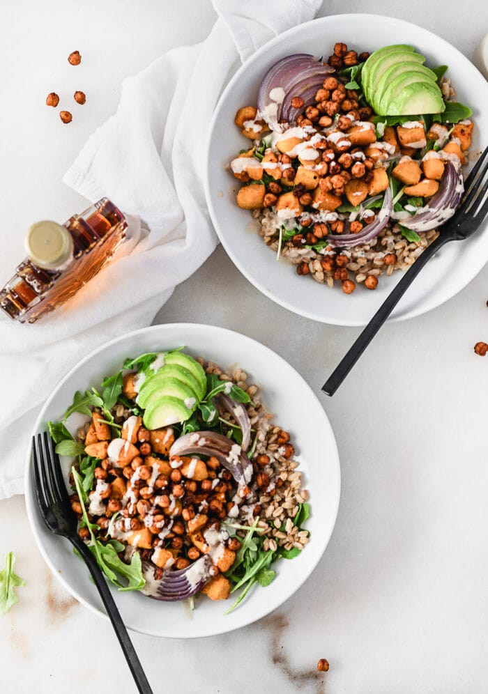 overhead view of two maple roasted chickpea grain bowls with roasted sweet potatoes, onions, arugula, and avocado with tahini dressing in white bowls with black forks in them, surrounded by a white napkin and a bottle of maple syrup.