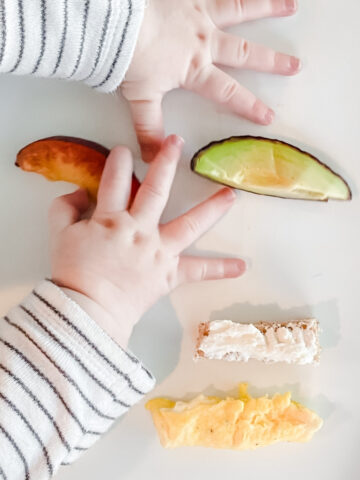 baby hands on a high chair tray with slices of avocado, plum, toast and eggs.