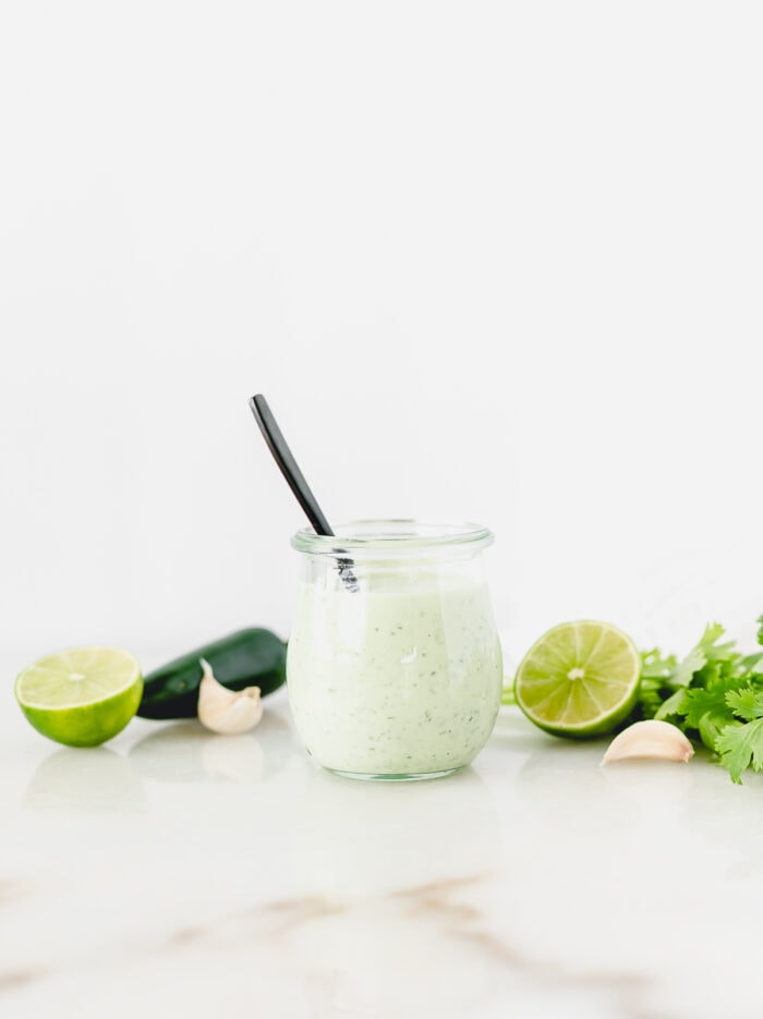 glass jar of creamy jalapeno lime dressing with a black spoon in it, surrounded by limes, cilantro and jalapenos.