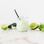 glass jar of creamy jalapeno lime dressing with a black spoon in it, surrounded by limes, cilantro and jalapenos.
