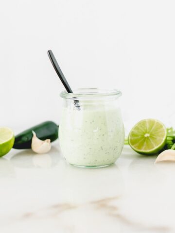 glass jar of creamy jalapeno lime dressing with a black spoon in it, surrounded by limes, cilantro and jalapenos.