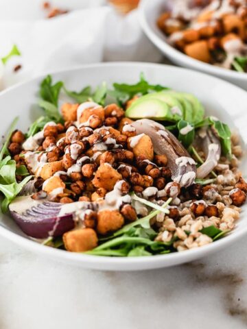 maple chili roasted chickpea grain bowl with roasted sweet potatoes, arugula, avocado and tahini dressing in a white bowl with another bowl in the background.