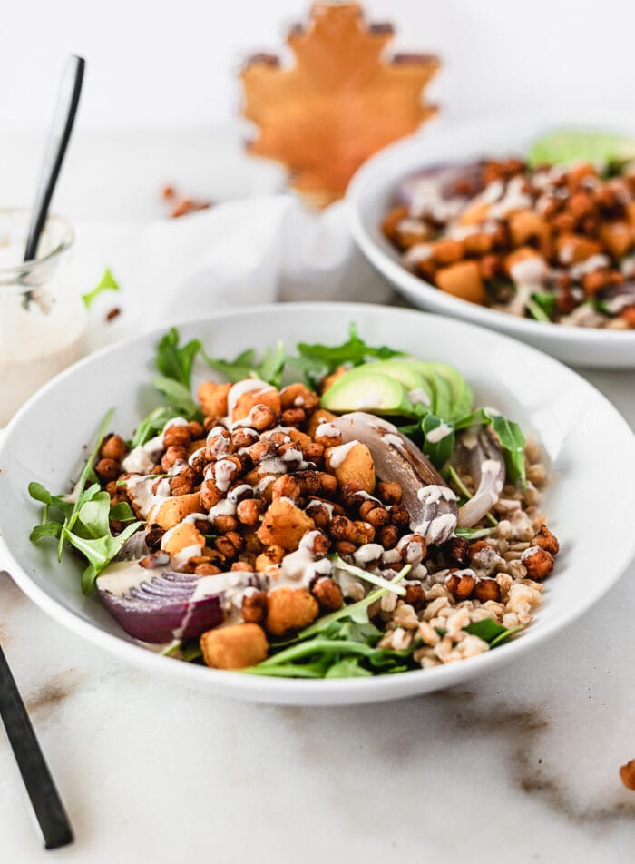 maple chili roasted chickpea grain bowl with roasted sweet potatoes, arugula, avocado and tahini dressing in a white bowl with another bowl and a bottle of maple syrup in the background.