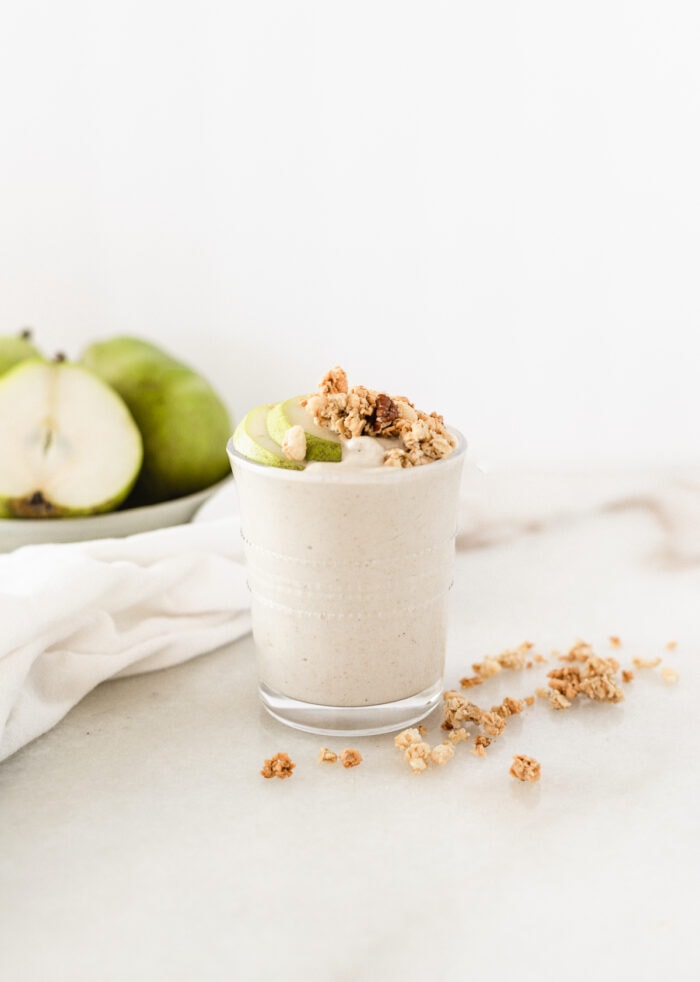 pear crisp smoothie in a glass topped with granola, with pears in the background.