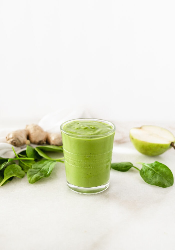 green smoothie in a glass surrounded by spinach, ginger root, and pears.