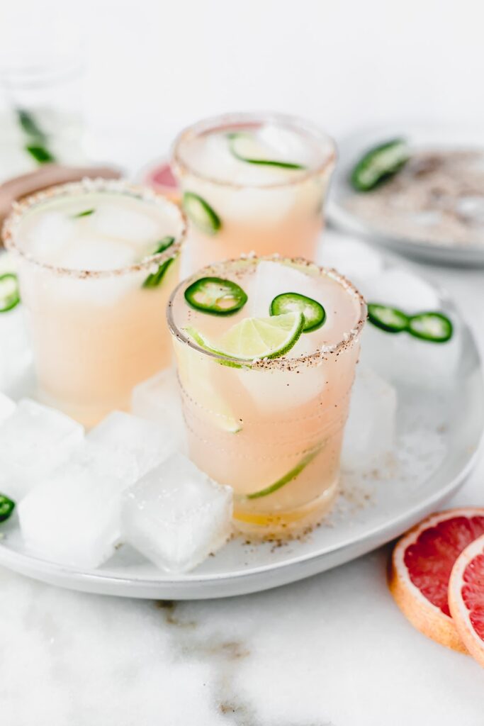three mezcal paloma cocktails with jalapeno slices and limes in them sitting on a grey plate with ice.