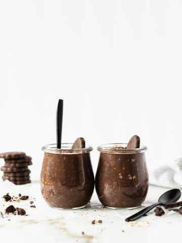 two glass jars of thin mint overnight oats with a black spoon in one, surrounded by thin mint cookies.
