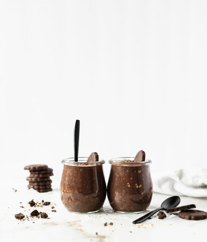 two glass jars of thin mint overnight oats with a black spoon in one, surrounded by thin mint cookies.