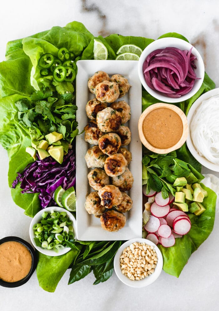 overhead view of a DIY meatball lettuce wrap tray with pork meatballs in the center surrounded by lettuce, herbs, jalapeño, pickled onions, peanut sauce, avocado and radishes. 