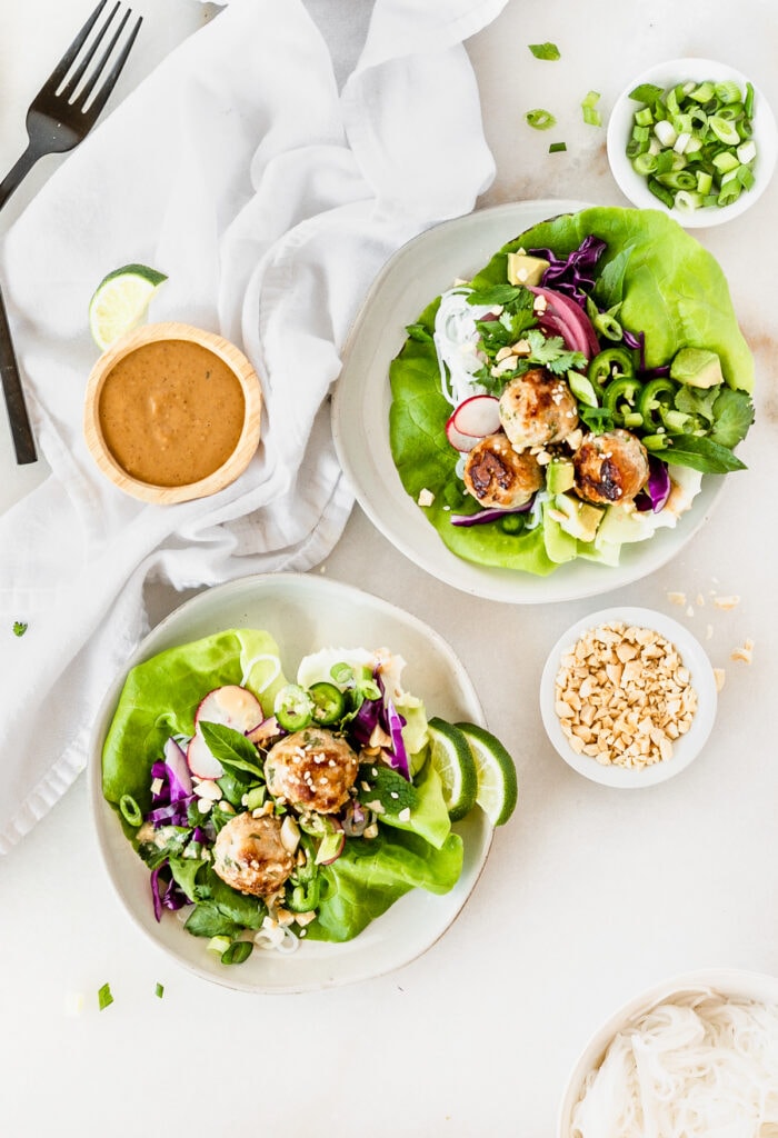 overhead view of two pork meatball lettuce wraps on small plates surrounded by small bowls of peanut dipping sauce, chopped peanuts, and green onions on a white background.