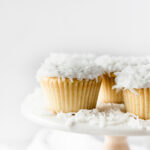 closeup of a coconut cupcake on a white cake stand with other coconut cupcakes.