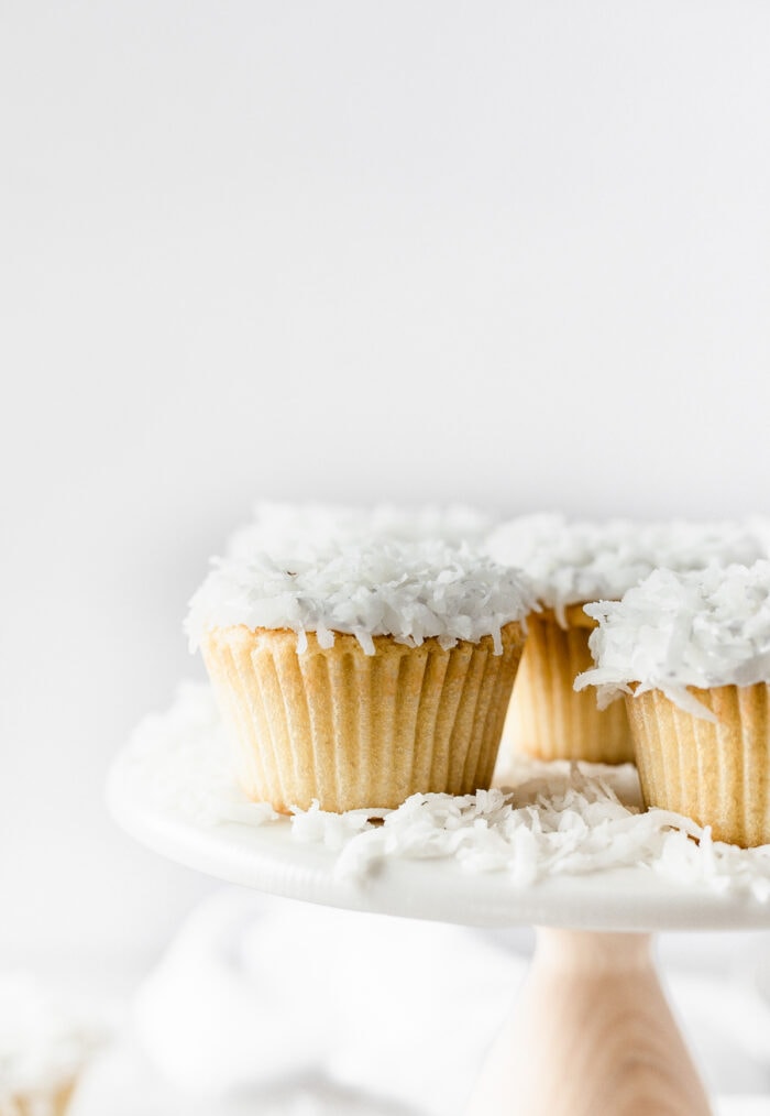closeup of a coconut cupcake on a white cake stand with other coconut cupcakes.