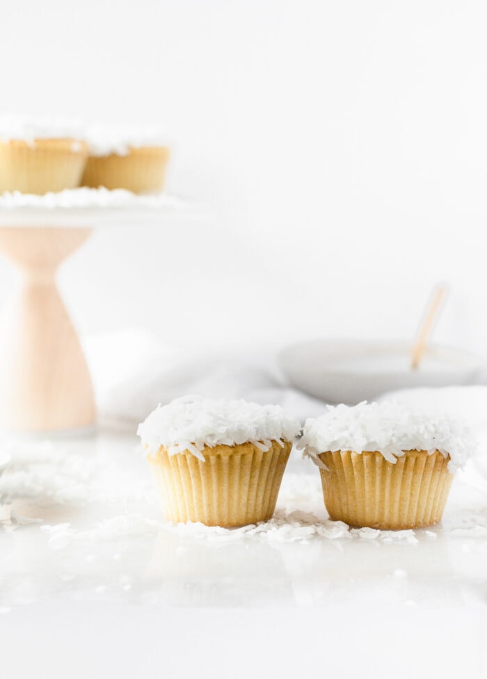 two coconut cupcakes with a cake stand of cupcakes and a bowl of frosting in the background.