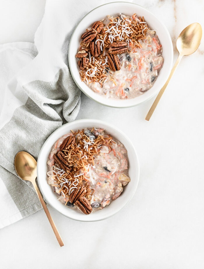 overhead view of two bowls of carrot cake overnight oats topped with toasted pecans and coconut flakes with a gold spoon beside each.