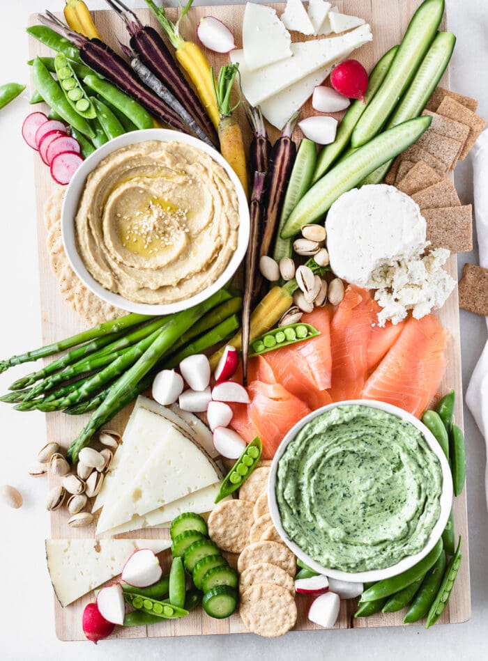 overhead view of a spring snack board with bowls of hummus and green dip, fresh veggies, smoked salmon and cheese.