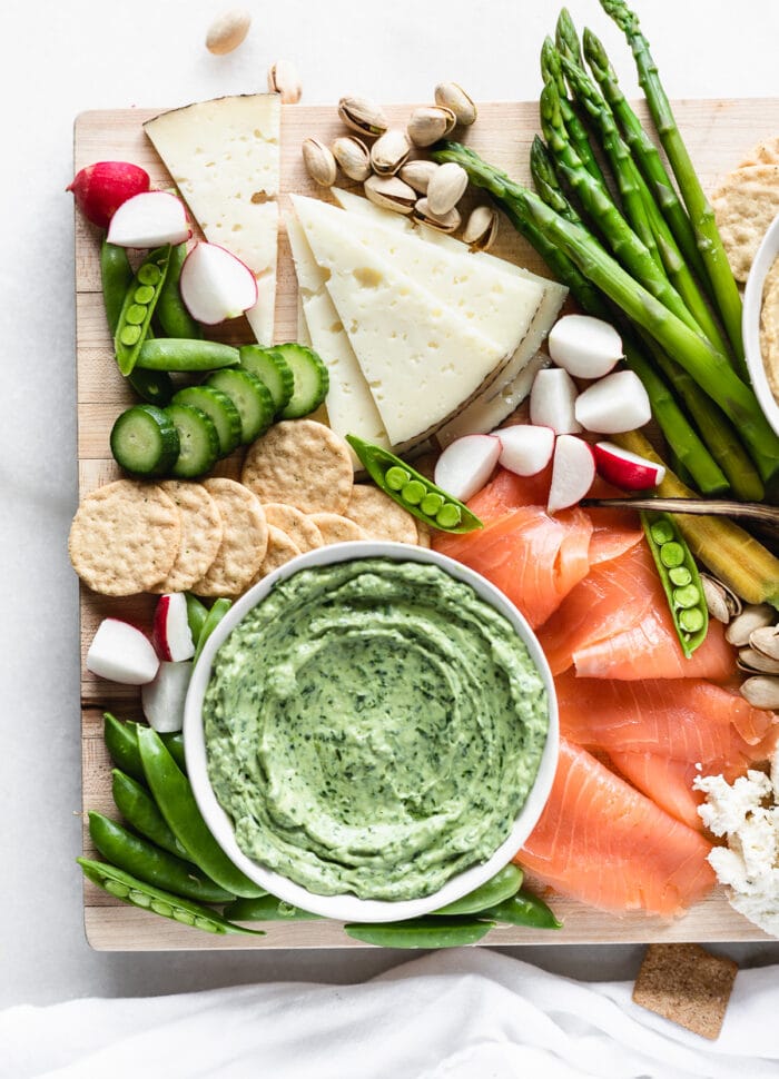 closeup of spring cheese board with a bowl of green goddess dip, white cheese, smoked salmon and fresh veggies.