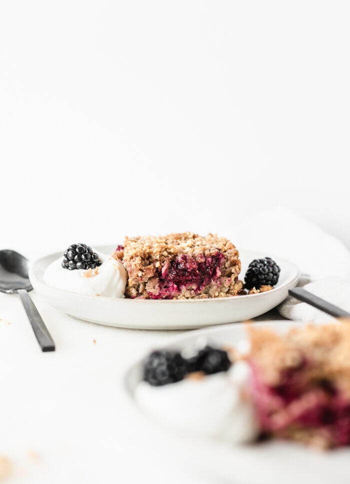 square of blackberry crumble baked oatmeal on a white plate with yogurt and blackberries with a black spoon beside it.