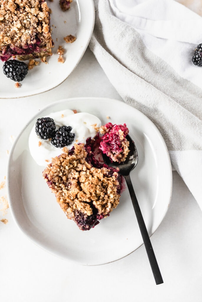 overhead view of a square of blackberry crumbled baked oatmeal on a plate with yogurt and berries with a bite sitting on a black spoon.