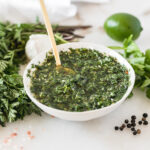 carrot top chimichurri in a small white bowl with a gold spoon in it surrounded by chimichurri ingredients.