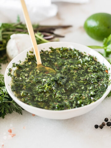 carrot top chimichurri in a small white bowl with a gold spoon in it surrounded by chimichurri ingredients.