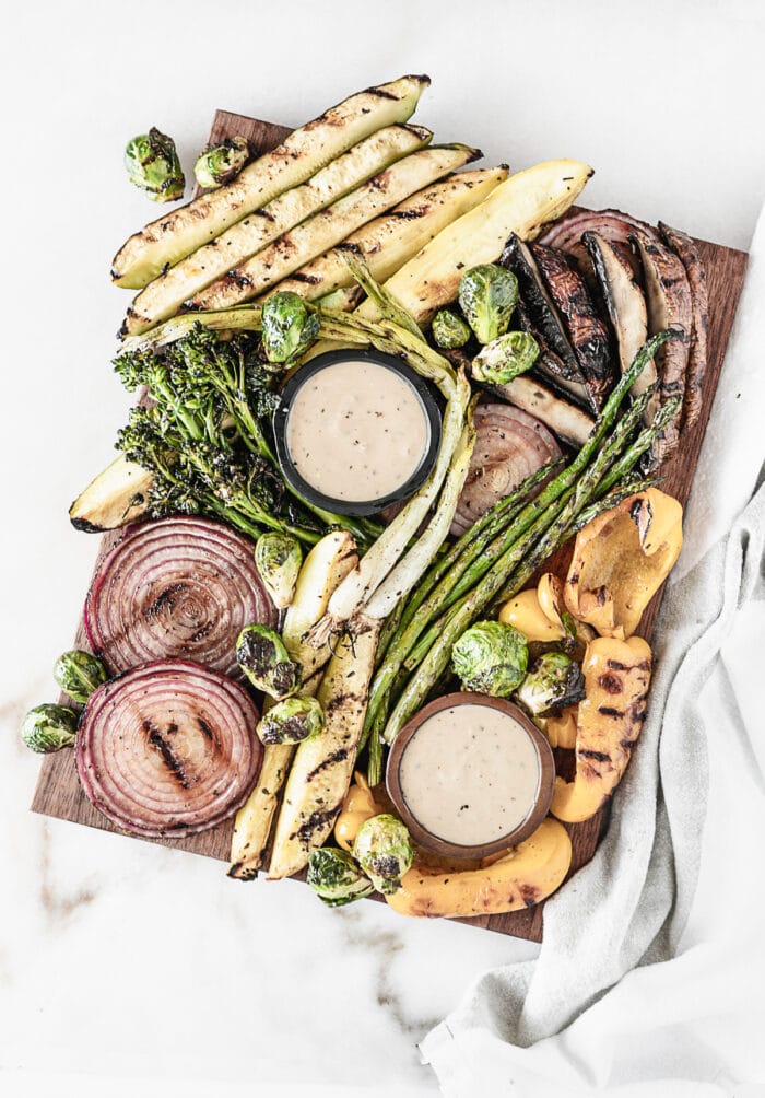 overhead view of various marinated grilled vegetables with small bowls of dipping sauce on a wooden board.