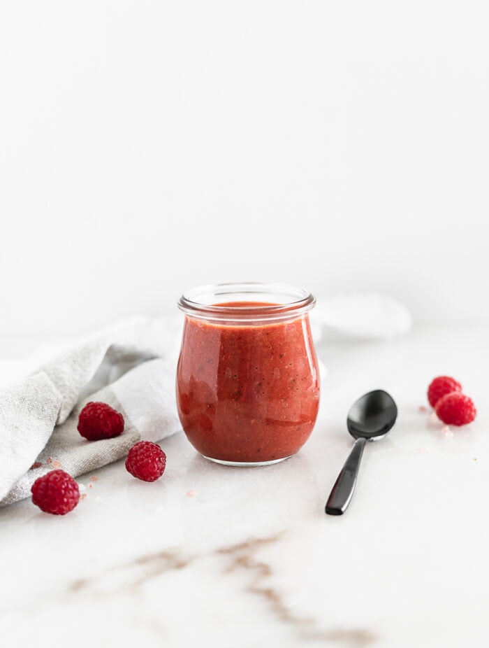 raspberry chipotle dressing in a glass jar with a black spoon beside it surrounded by fresh raspberries.