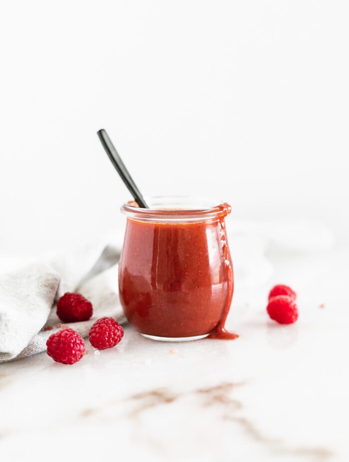 raspberry chipotle dressing in a glass jar with a black spoon in it surrounded by fresh raspberries.