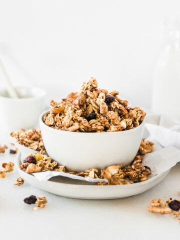 sourdough discard granola in a white bowl on top of a plate with granola clusters all around it.