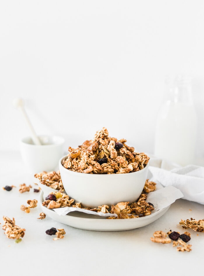 sourdough discard granola in a white bowl on top of a plate with granola clusters all around it.