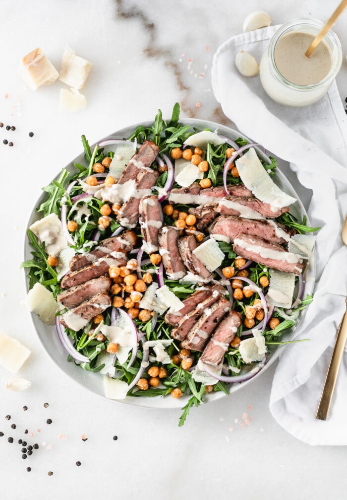 overhead view of steak and arugula salad with roasted chickpeas and tahini dressing on a plate with a jar of dressing beside it.