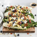 grilled romaine halves topped with corn, goat cheese, blueberries and dressing on a wood cutting board.