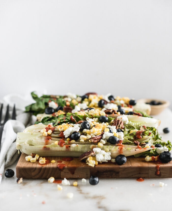 grilled romaine halves topped with corn, goat cheese, blueberries and dressing on a wood cutting board.