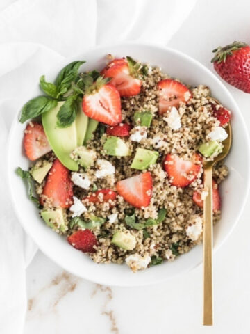overhead view of strawberry avocado quinoa salad in a white bowl with a gold spoon in it.