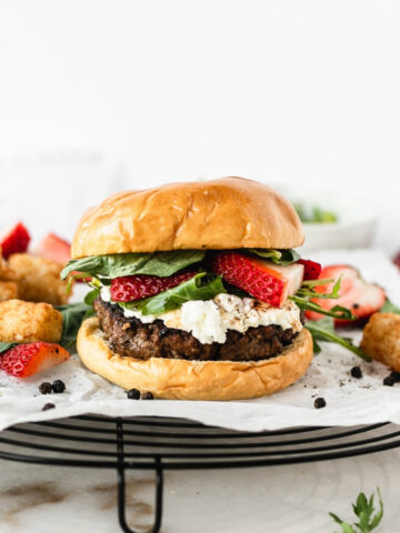strawberry basil goat cheese burger on a black wire rack topped with parchment with tater tots and strawberries in the background.