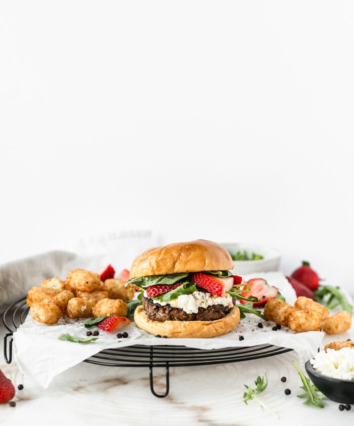 strawberry basil goat cheese burger on a black wire rack topped with parchment with tater tots and strawberries in the background.