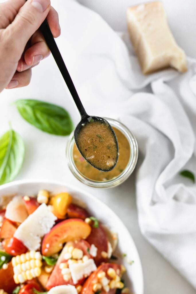 overhead view of a spoon of miso dijon dressing over the jar of dressing with basil, a bowl of peach salad, and a white napkin around it. 