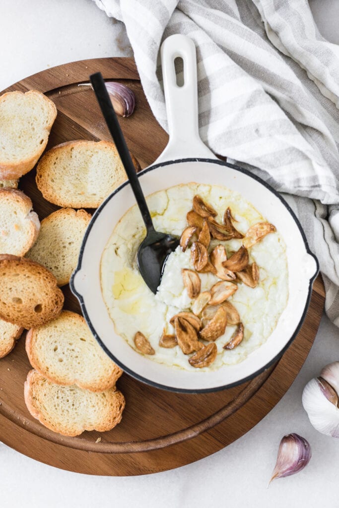 overhead view of a white skillet of hot goat cheese dip topped with crispy garlic slices with a black spoon in it surrounded by crostini and a grey and white linen..