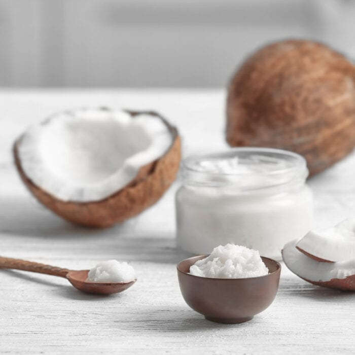 small brown bowl of coconut oil with a spoon and jar of coconut oil and a coconut half in the background.