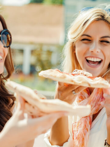 two women smiling and holding slices of pizza.