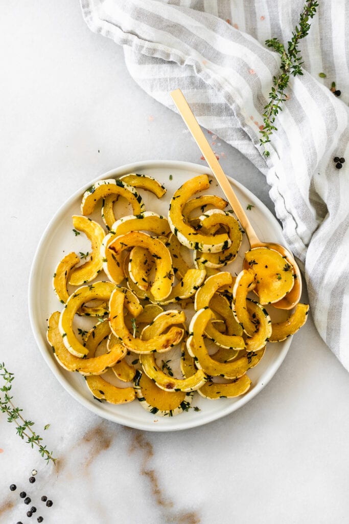 overhead view of roasted delicata squash half moons on a white plate with a gold spoon surrounded by thyme sprigs and a grey and white linen..