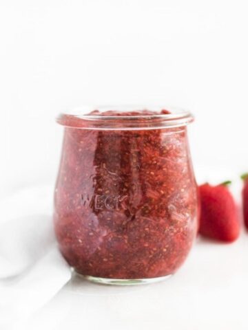 glass jar of strawberry rhubarb chia jam with strawberries behind it.