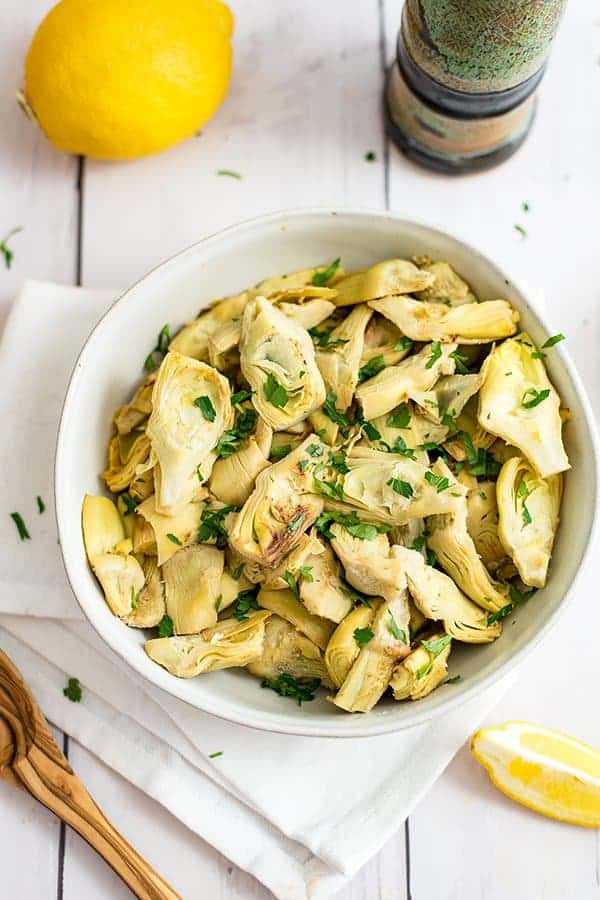 lemon artichoke in a bowl topped with fresh herbs surrounded by lemons, olive oil and a wooden spoon.