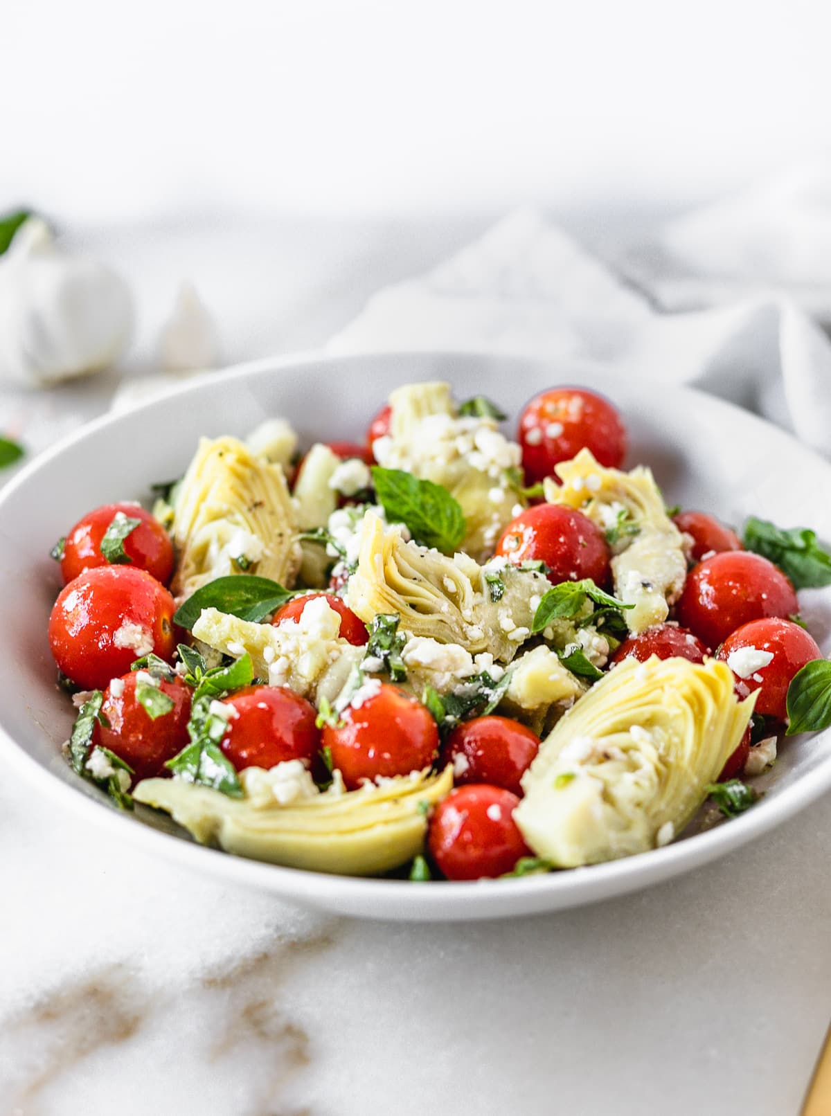 closeup of marinated artichoke salad with tomatoes herbs and feta in a white bowl.