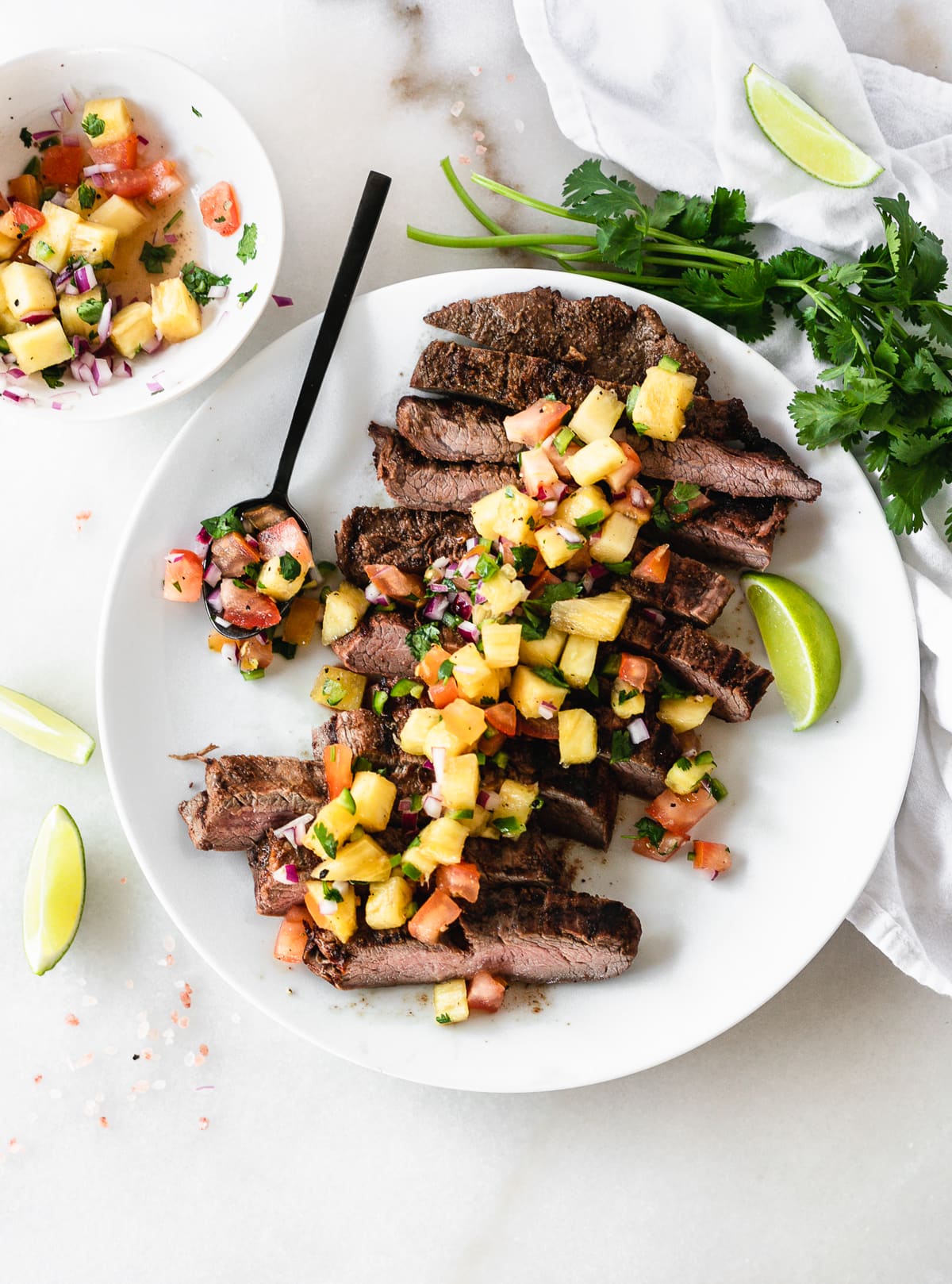 overhead view of sliced chipotle flank steak topped with pineapple salsa on a white plate with a spoon of salsa and lime wedges.